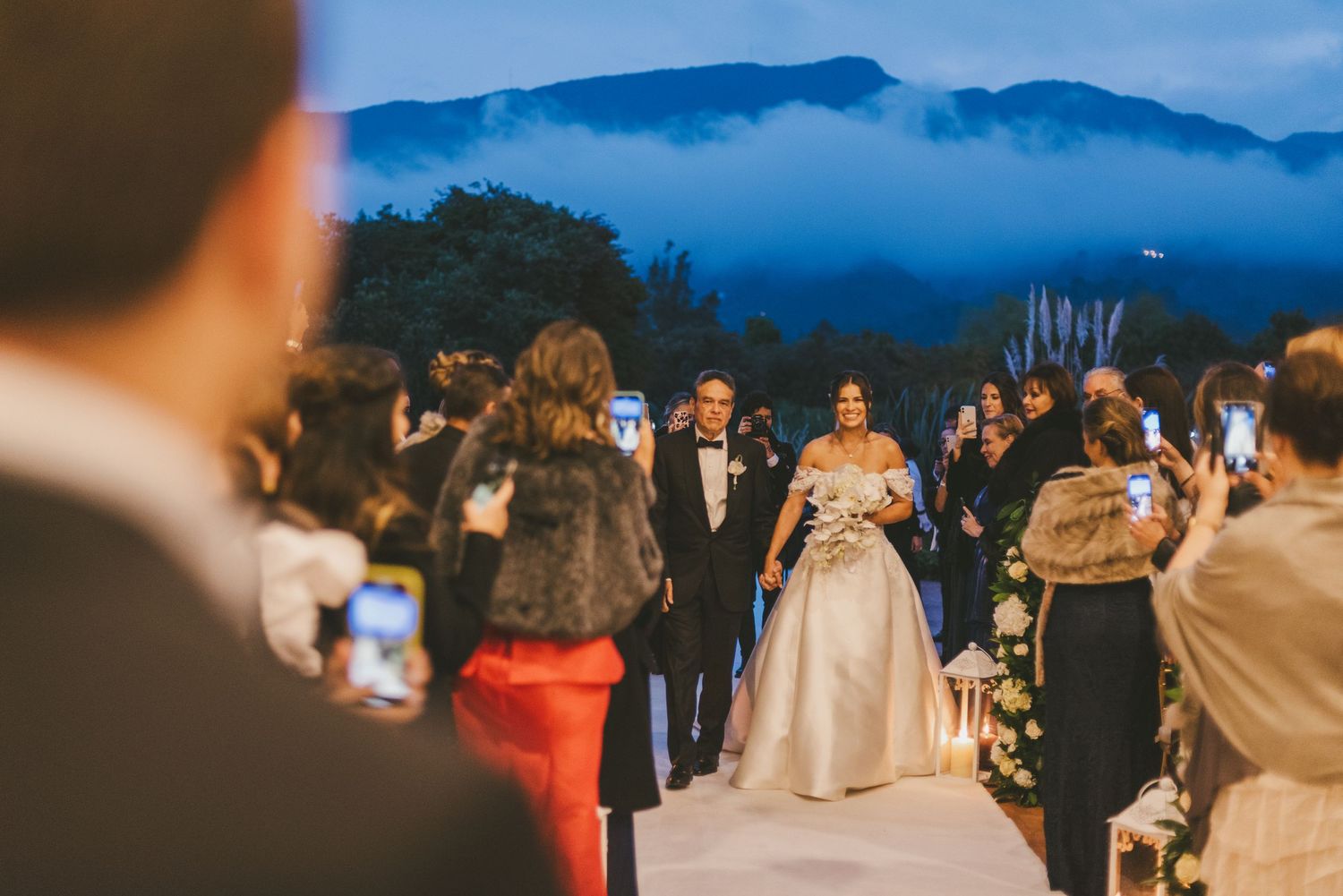 An outdoor evening wedding ceremony takes place against a backdrop of misty blue mountains at dusk.