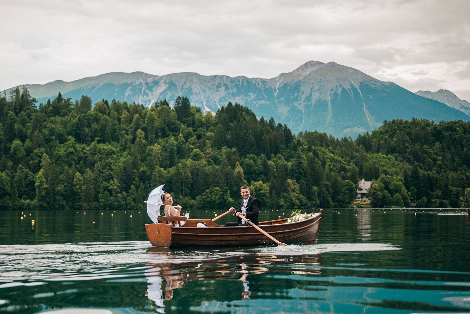 A wooden boat with people rowing on a serene lake surrounded by forested mountains under cloudy skies.