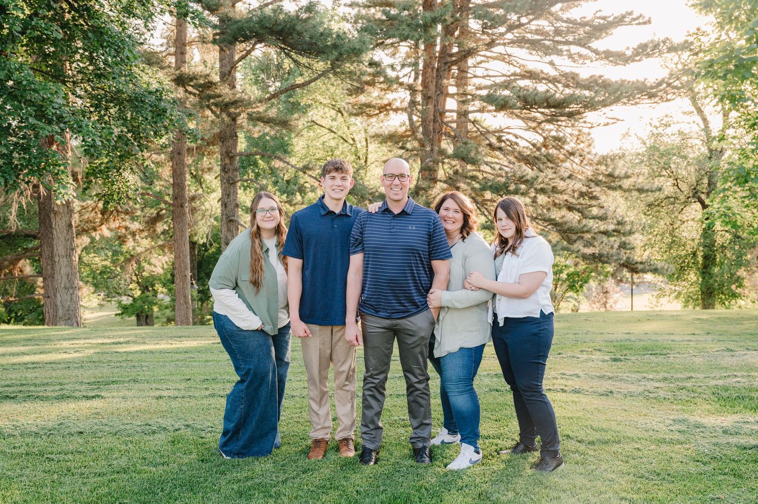 A group of people standing together outdoors in a sunlit park with tall pine trees in the background.