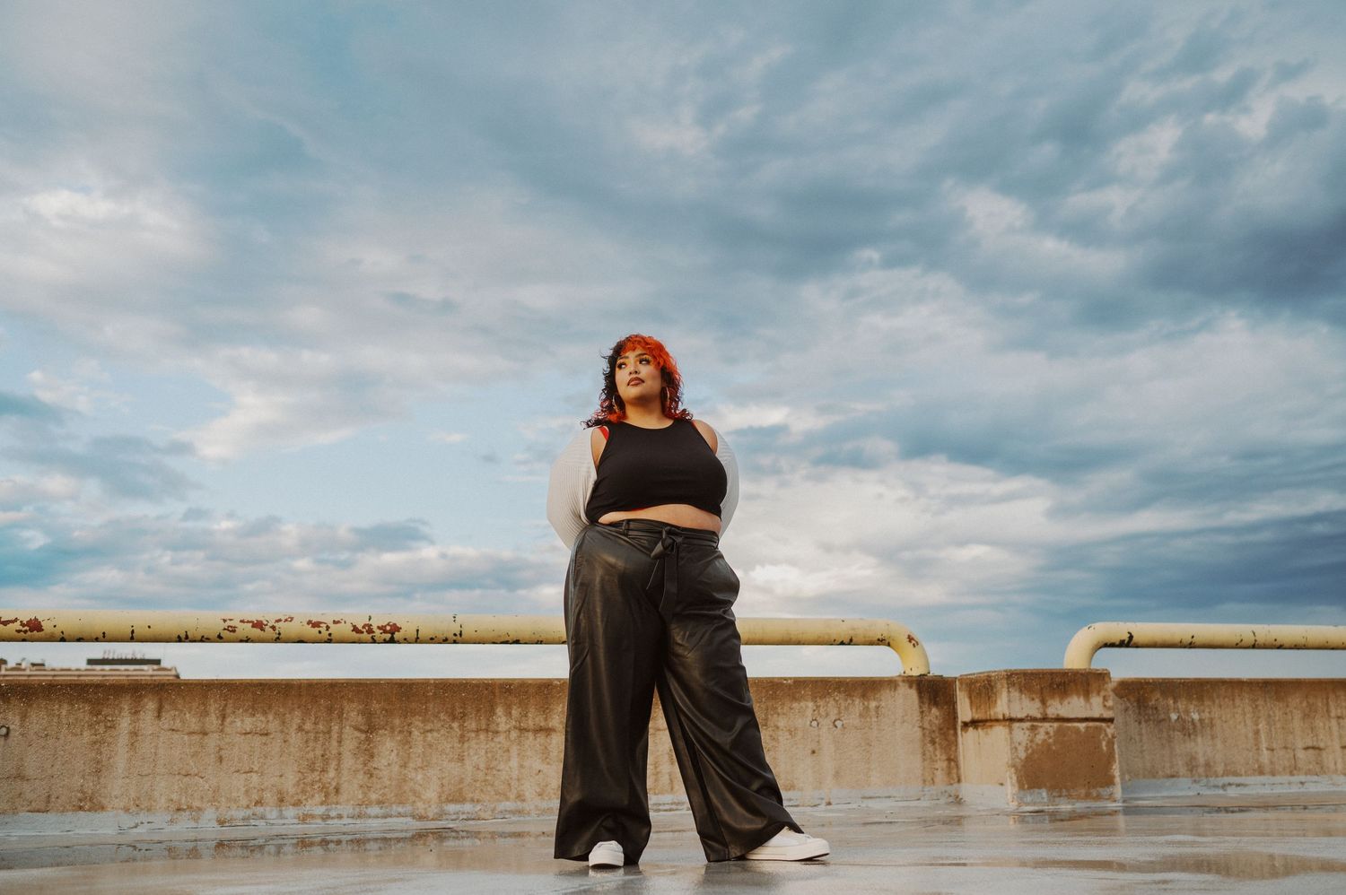 A figure in wide-leg pants and crop top stands on a rooftop against dramatic cloudy skies at sunset.