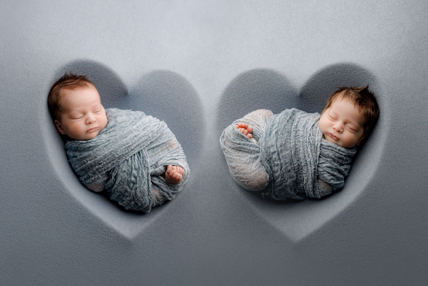 Newborn twins swaddled in grey positioned in heart-shaped frames against grey background.