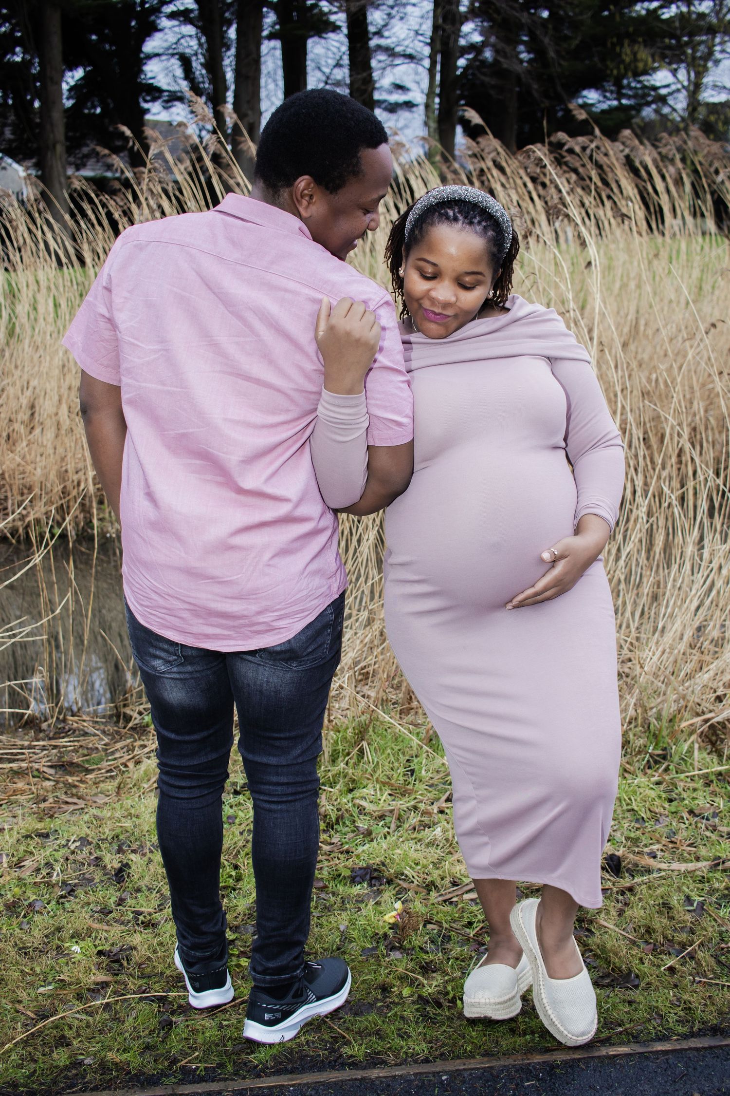 A couple in matching pink outfits share an intimate moment together in a field of tall grass during a maternity photoshoot.