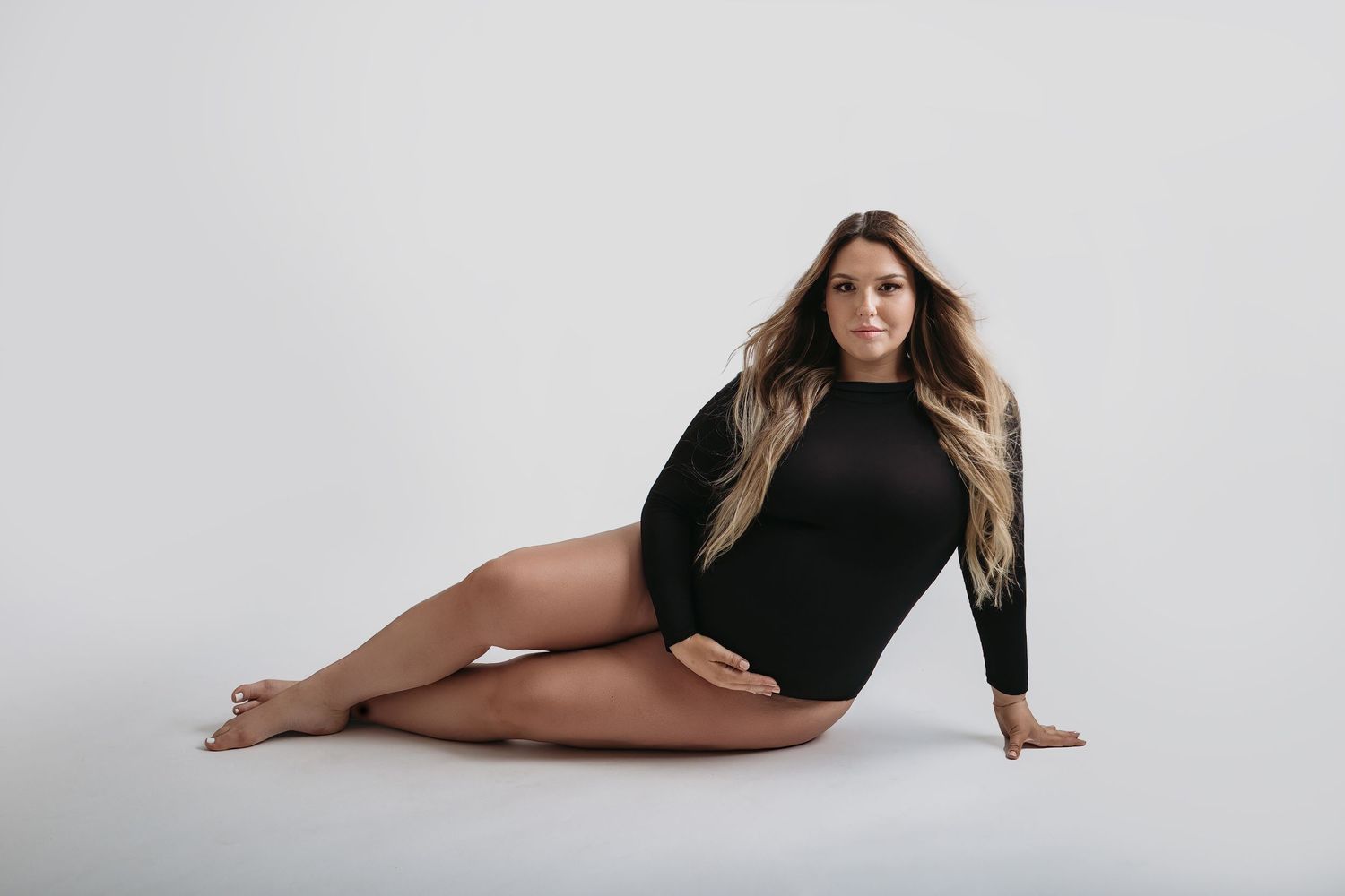 A pregnant woman in a black long sleeve bodysuit poses gracefully on a white studio floor for tampa maternity photo shoot