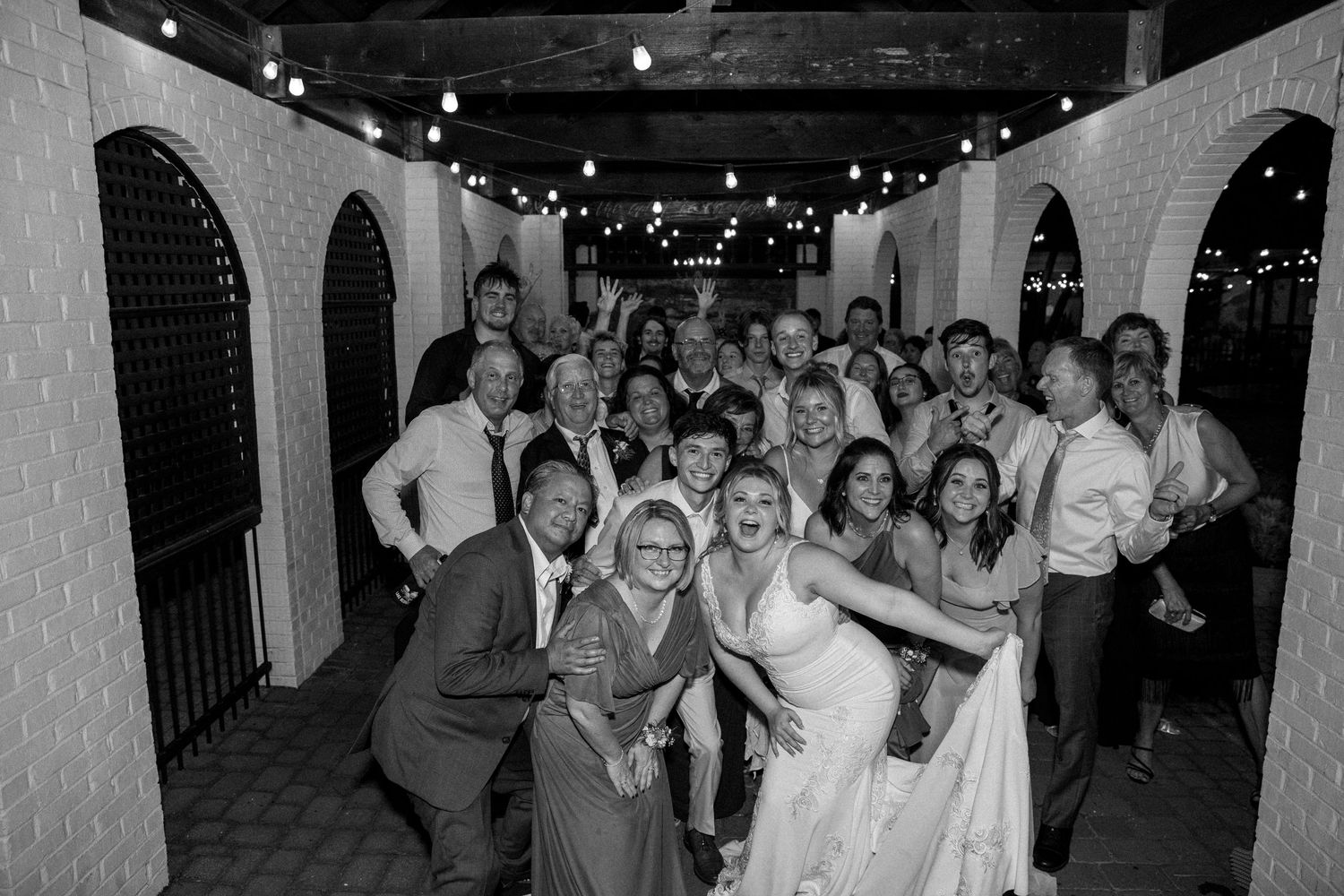 Wedding guests and party members gather for a fun group photo in an archway-lined venue.