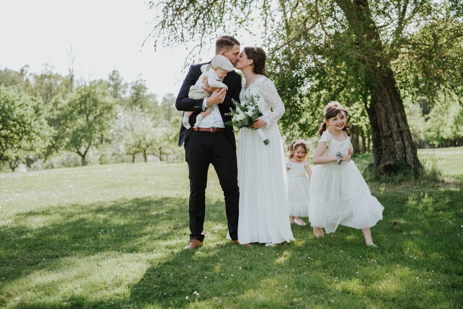 A wedding scene with a couple kissing while a flower girl in white stands nearby under a tree.
