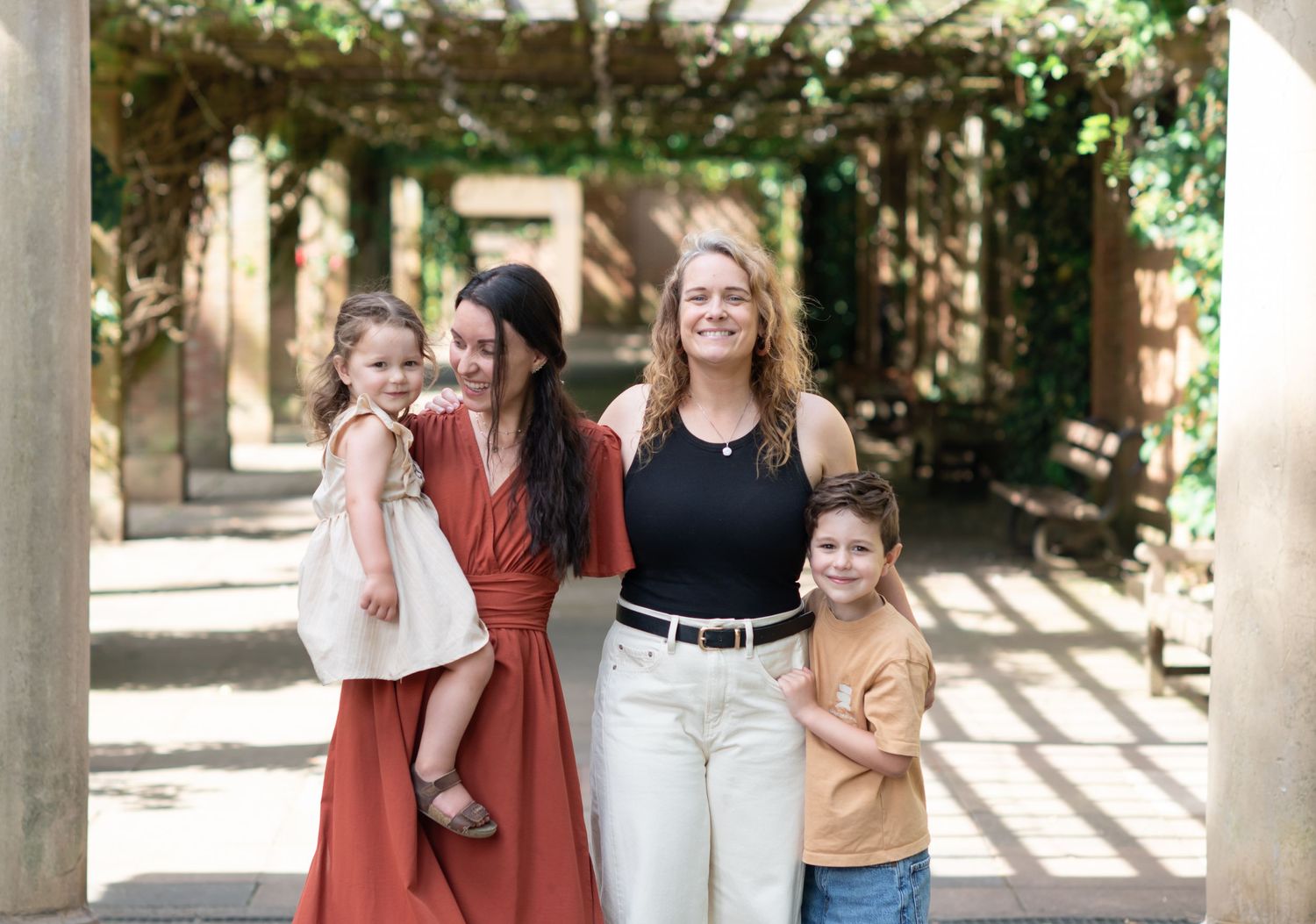 A family stand together beneath an outdoor pergola with dappled sunlight casting shadows on the walkway.