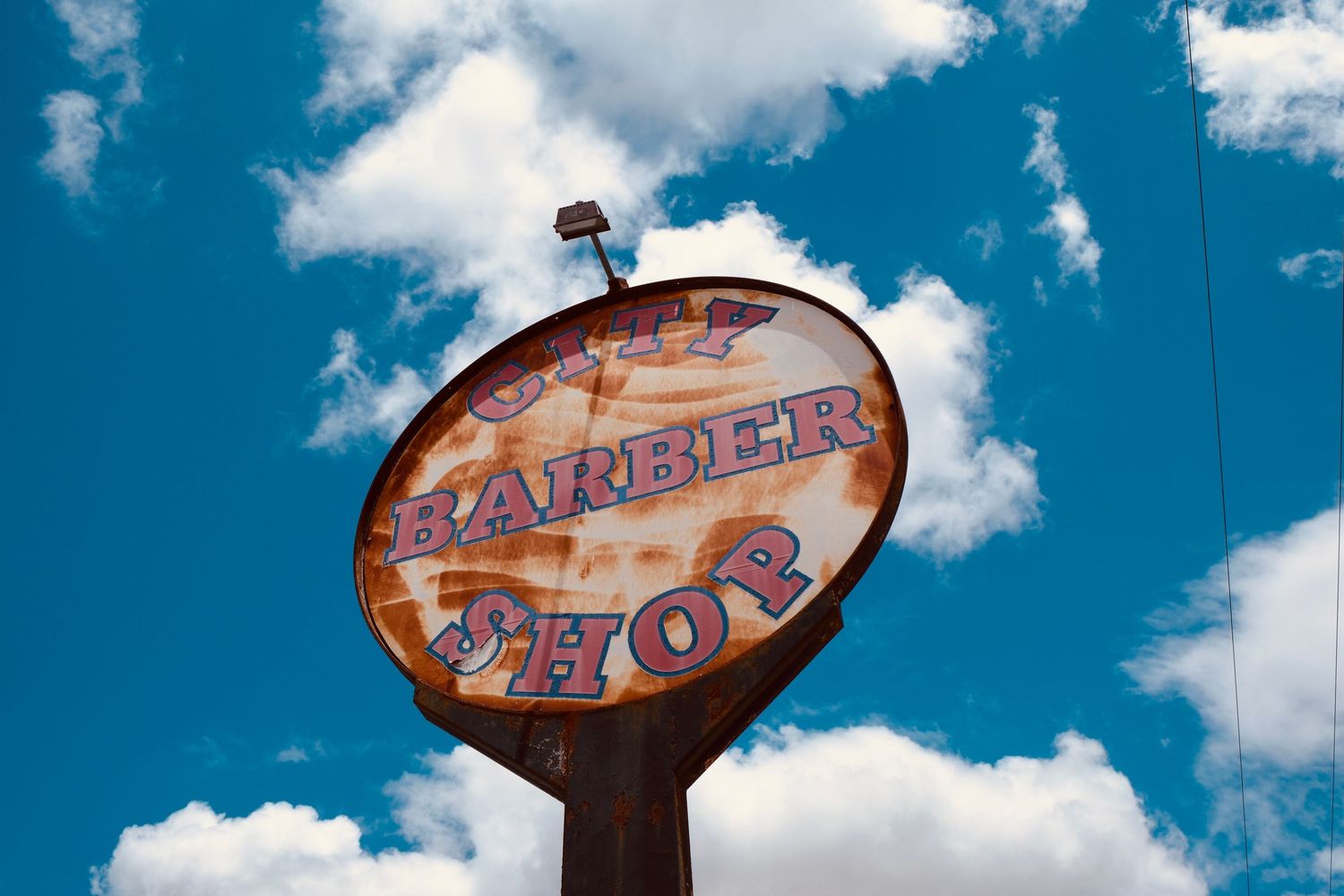 Vintage rusty barber shop sign against a bright blue sky with white clouds in a small town.