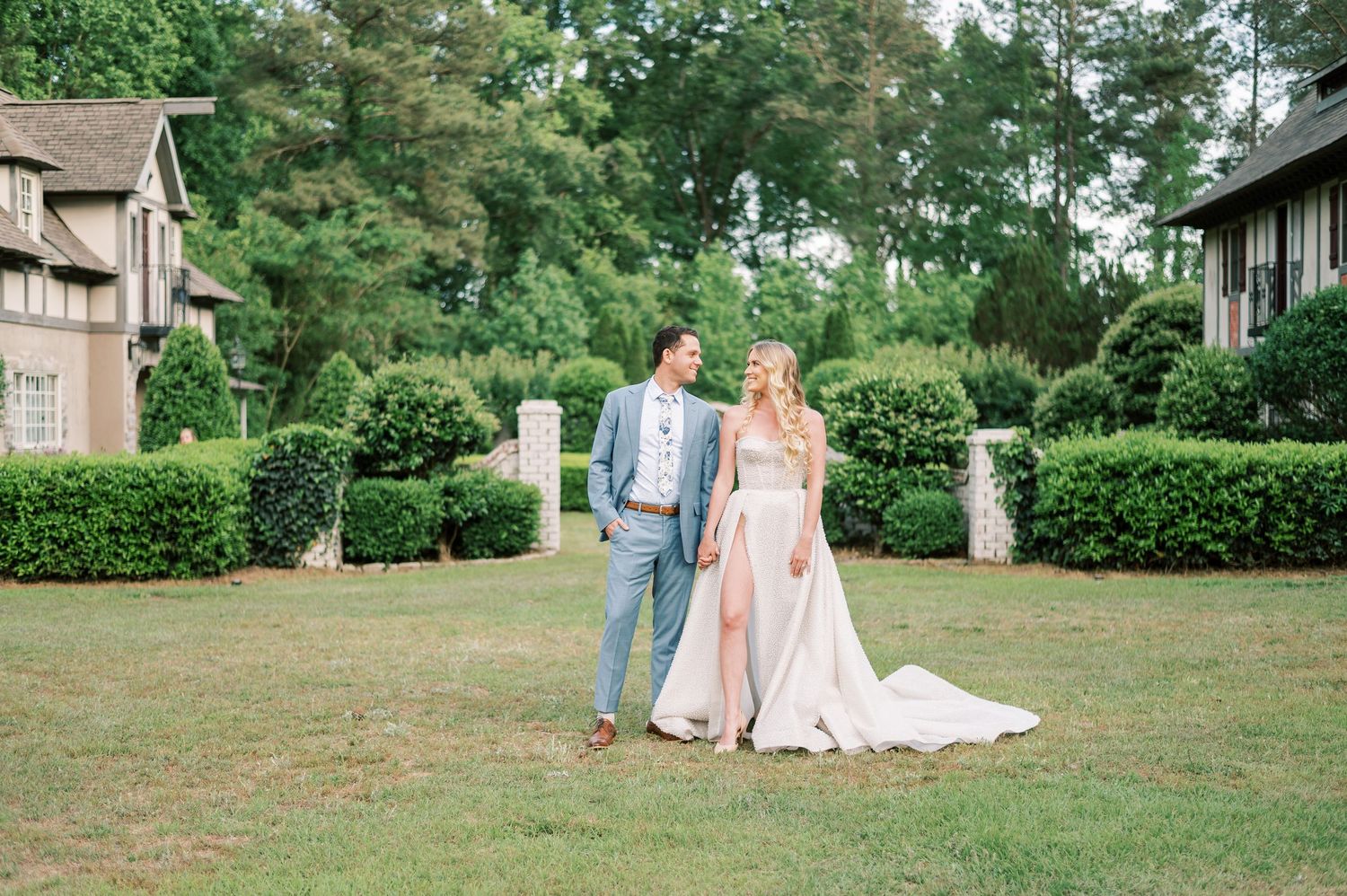A couple stands elegantly on a manicured lawn surrounded by hedges and stately homes in formal wedding attire.