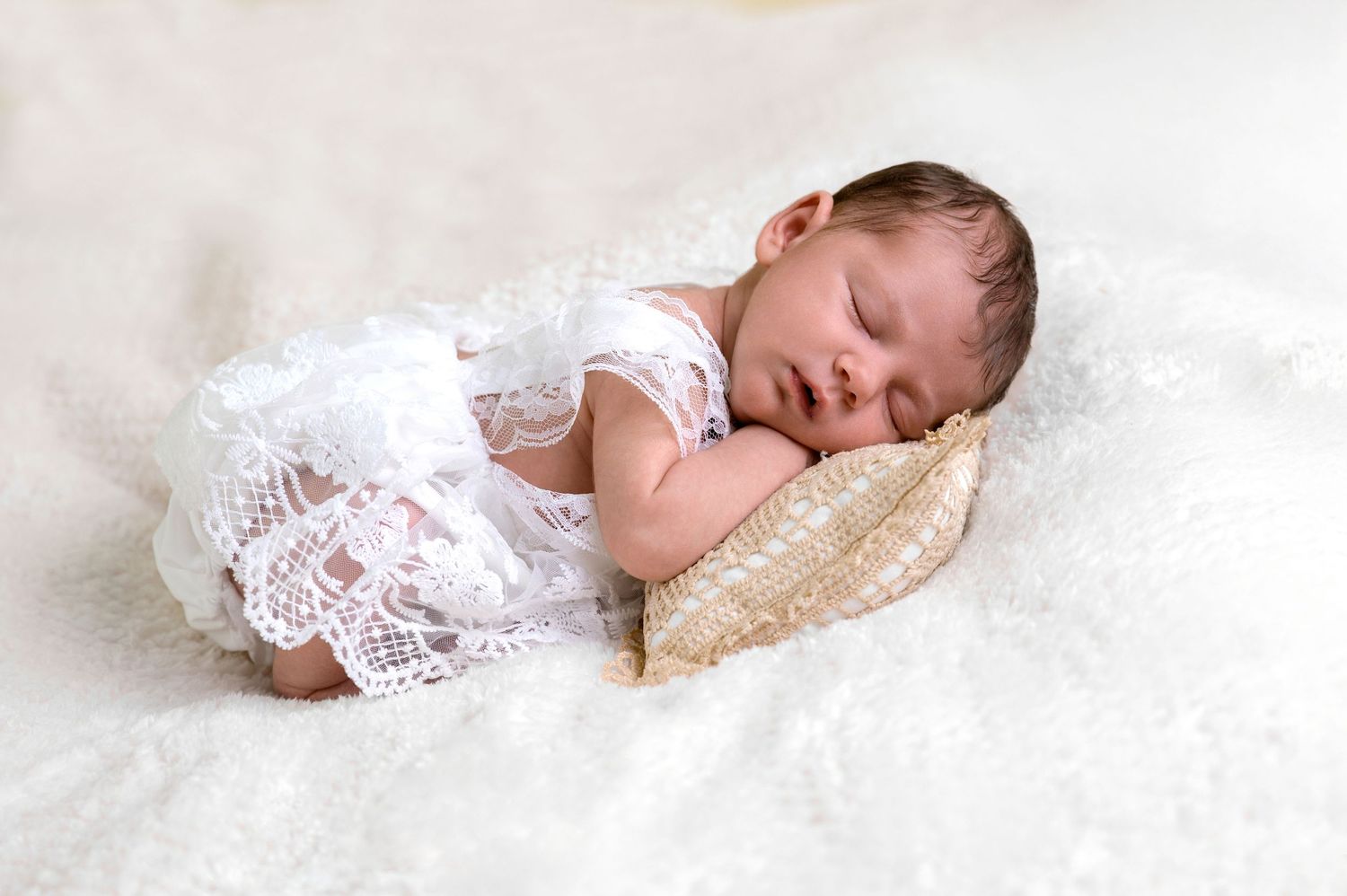 Sleeping newborn wearing white lace dress rests peacefully on cream-colored pillow.