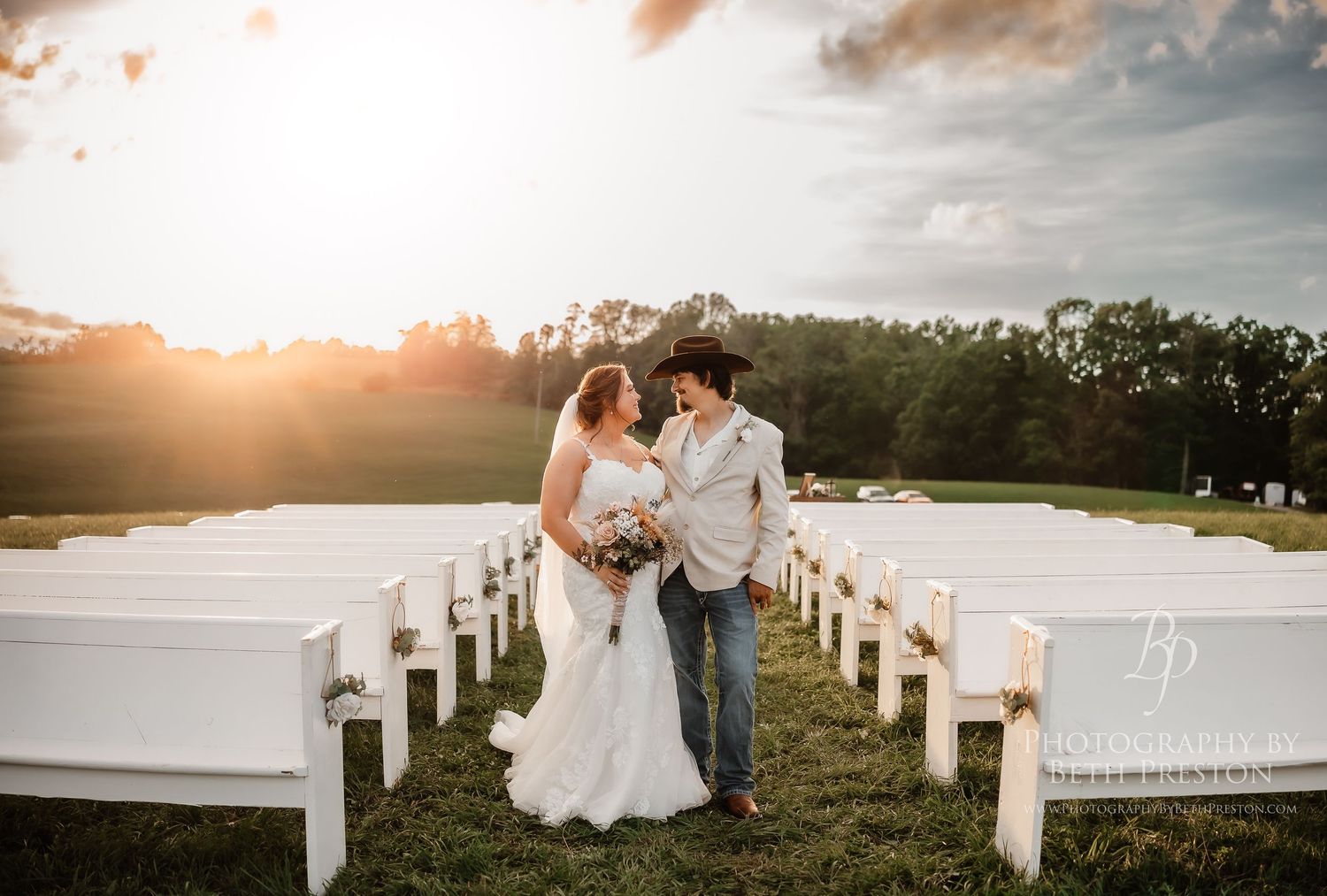 A couple embraces at sunset in front of white wooden chairs set up for an outdoor wedding ceremony.