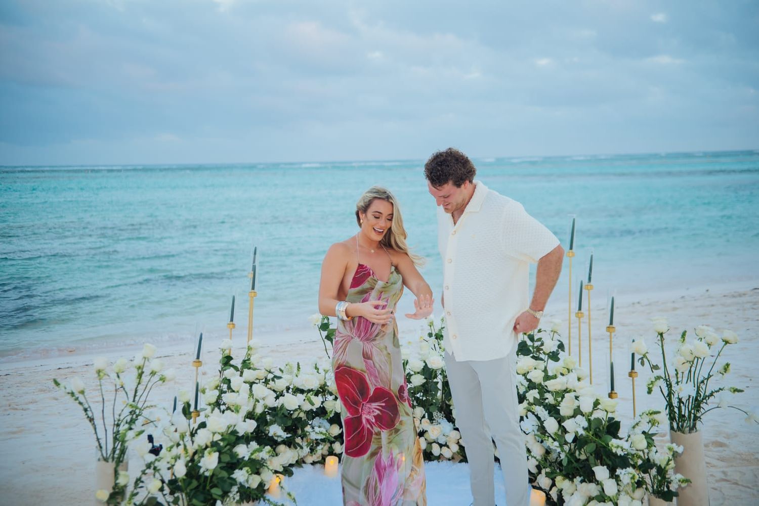 Romantic beach proposal setup with white floral arrangements and candles along turquoise ocean waters.