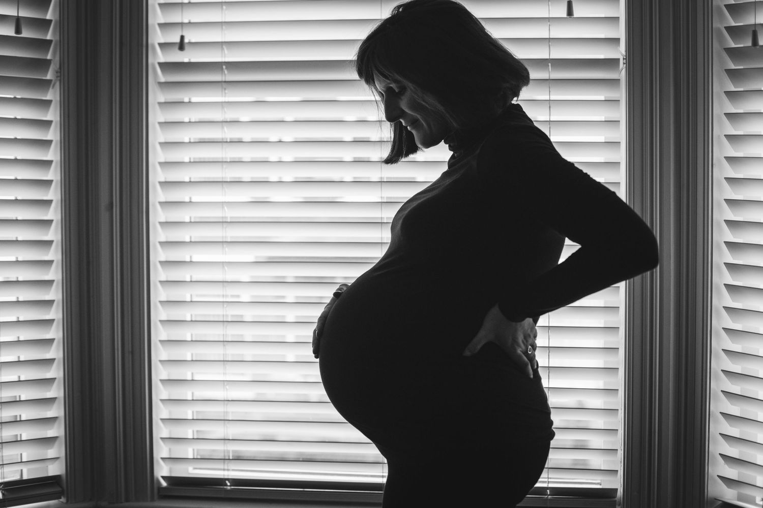 Black and white silhouette of an expectant mother standing by a window with venetian blinds in the background.