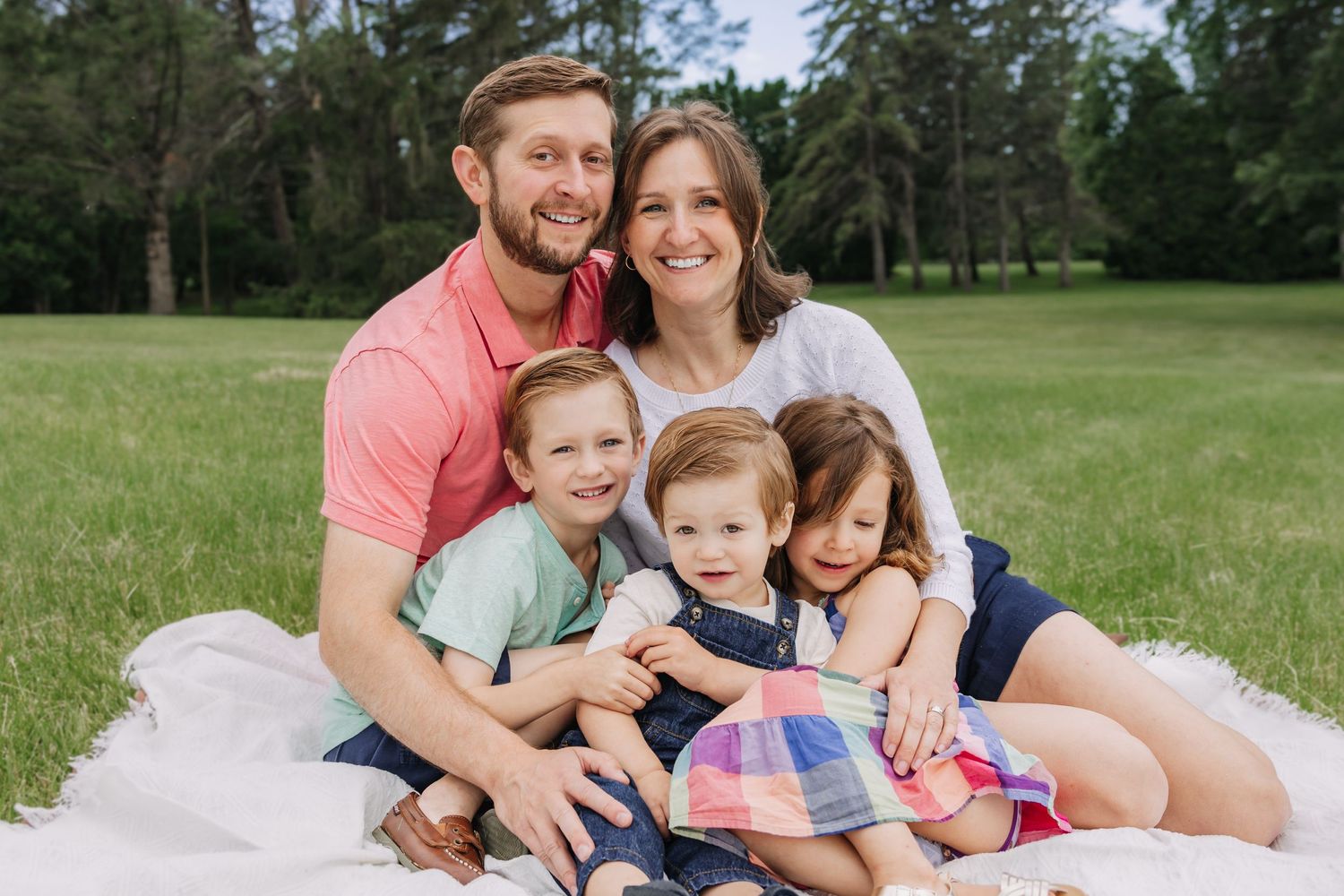 Family sitting together on white blanket in sunny field surrounded by pine trees.