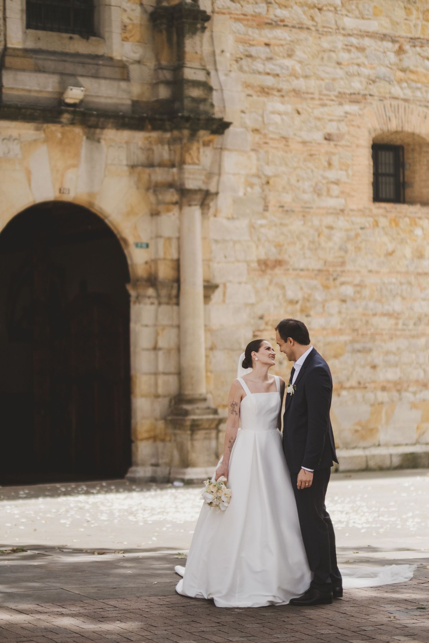 A romantic wedding couple share an intimate moment outside a historic stone building with arched doorways.