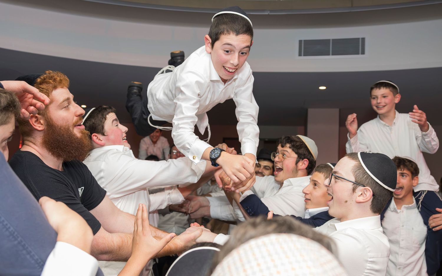 A group celebrates at an indoor celebration by lifting and supporting someone in white dress attire during a festive gathering.