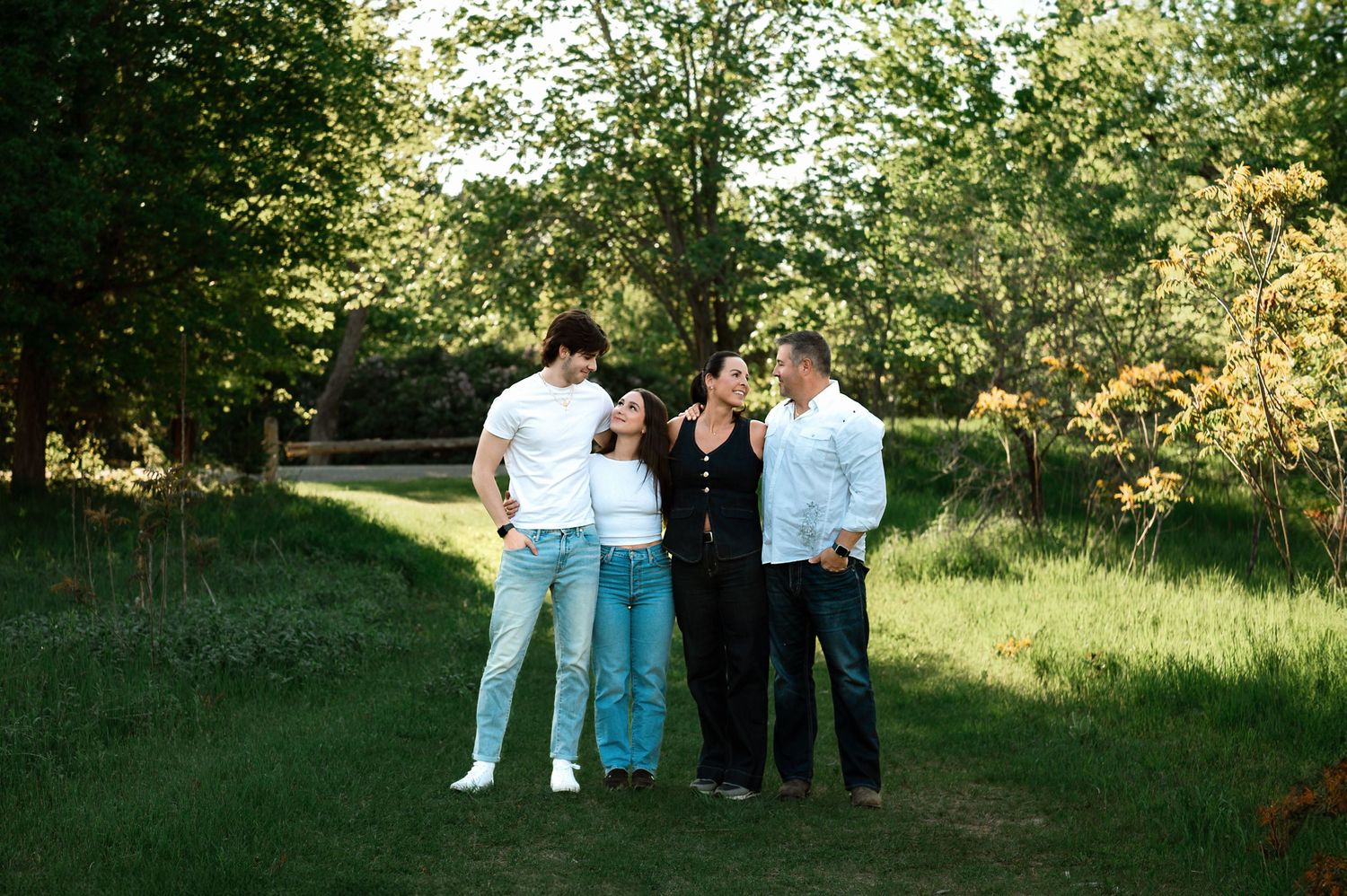 Four people stand together outdoors on a grassy path surrounded by green trees on a sunny day.