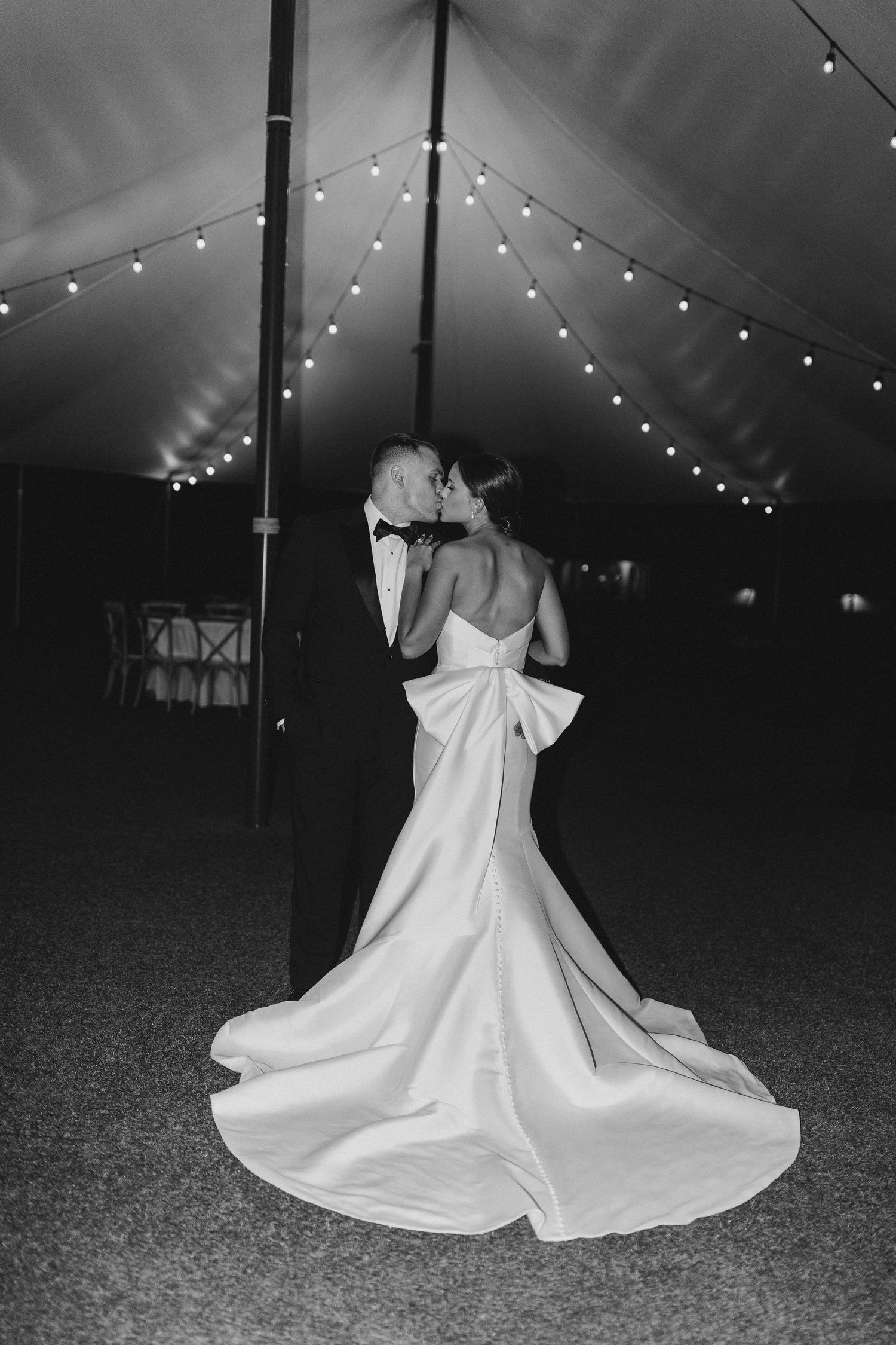Black and white photograph of a couple embracing under a wedding tent with string lights creating a romantic atmosphere.