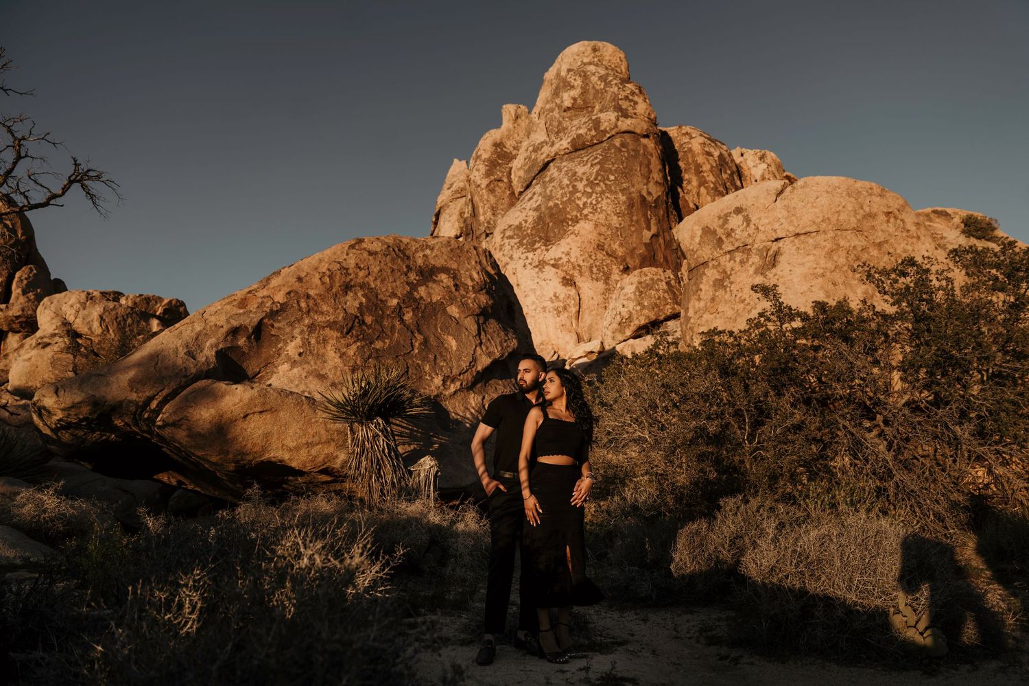 Two figures stand in silhouette against large golden rock formations at sunset in Joshua Tree National Park.