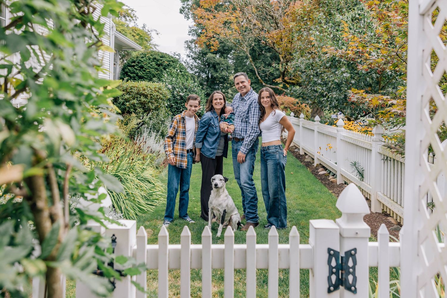 Happy family of four with a white dog posing in a lush backyard garden behind a white picket fence.