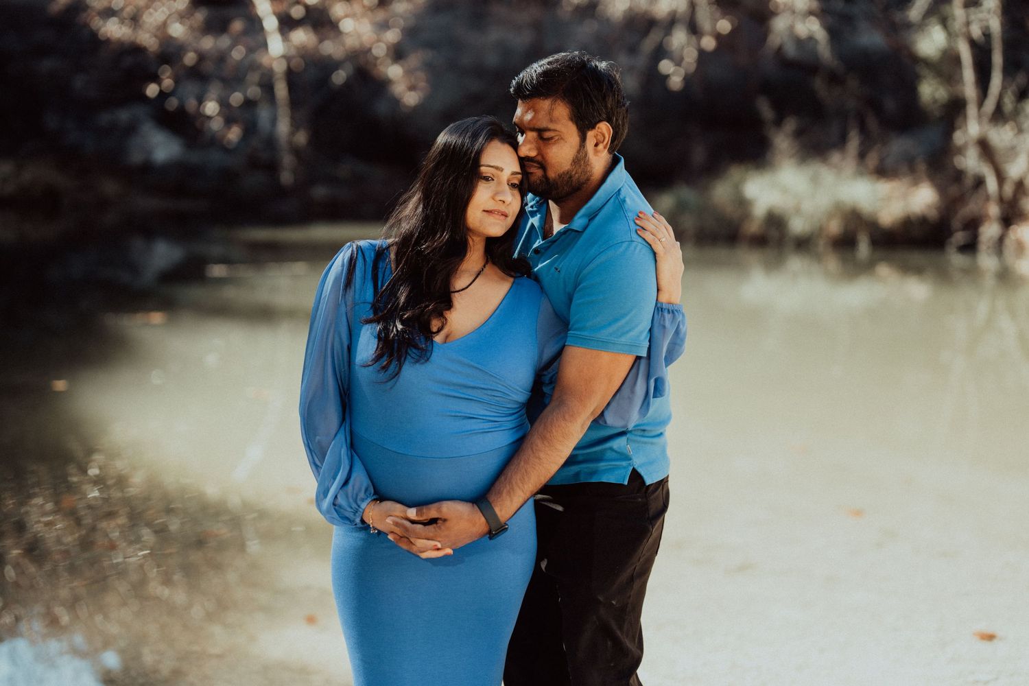 A maternity photo session by a lake with a couple in coordinating blue outfits.