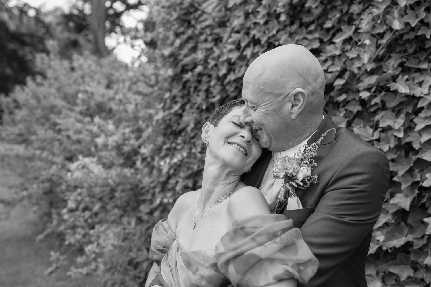Black and white photo of a loving mature couple sharing an intimate moment against an ivy-covered wall backdrop.