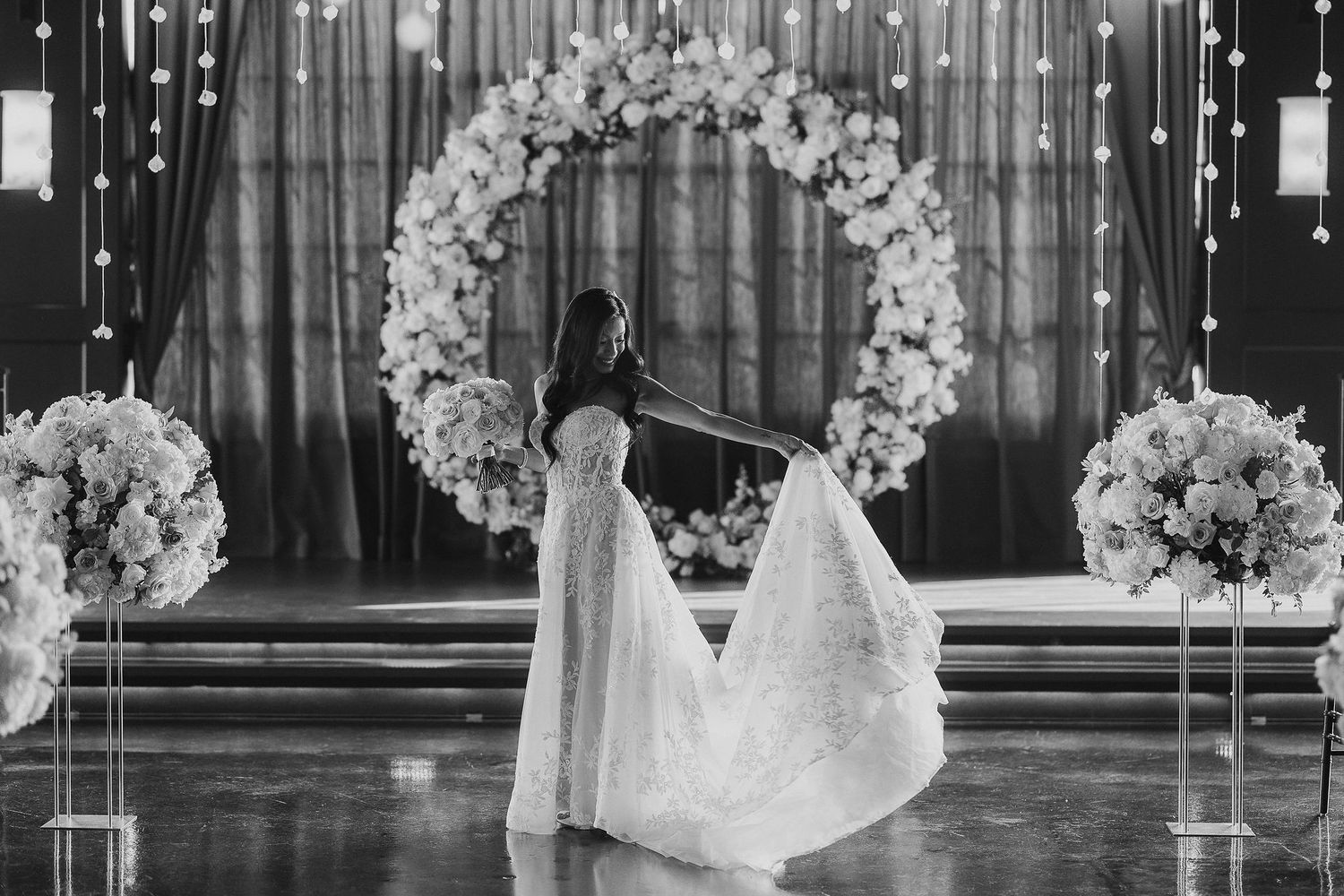 Black and white photo of bride twirling dress under floral arch with string lights.