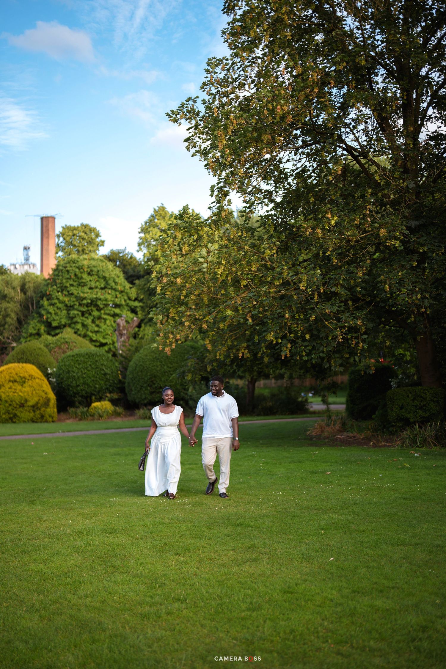 Couple in white outfits holding hands while walking on lush green lawn in a scenic park on a sunny day.