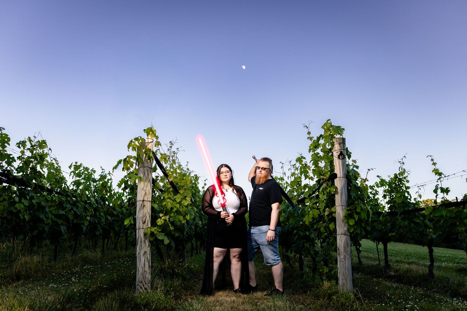 Couple holding lightsabers in a vineyard at dusk with lush green vines and a clear blue sky.
