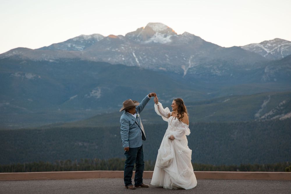Couple dancing at mountain overlook, bride in white gown, groom in hat, snowy peaks backdrop at sunset.