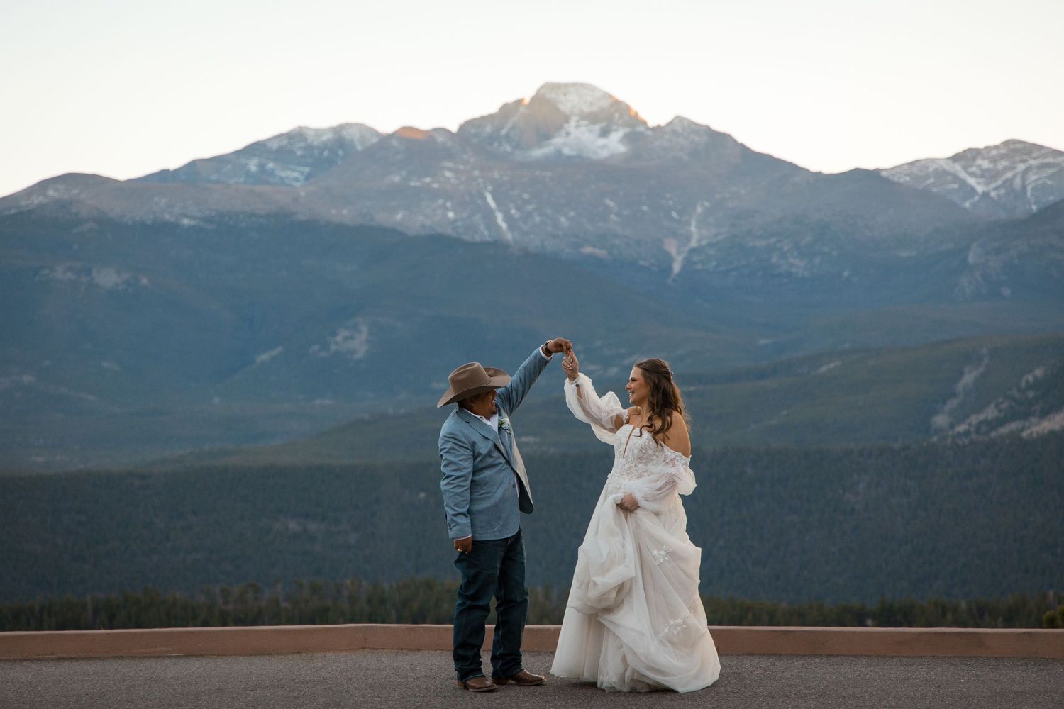 Couple dancing at mountain overlook, bride in white gown, groom in hat, snowy peaks backdrop at sunset.