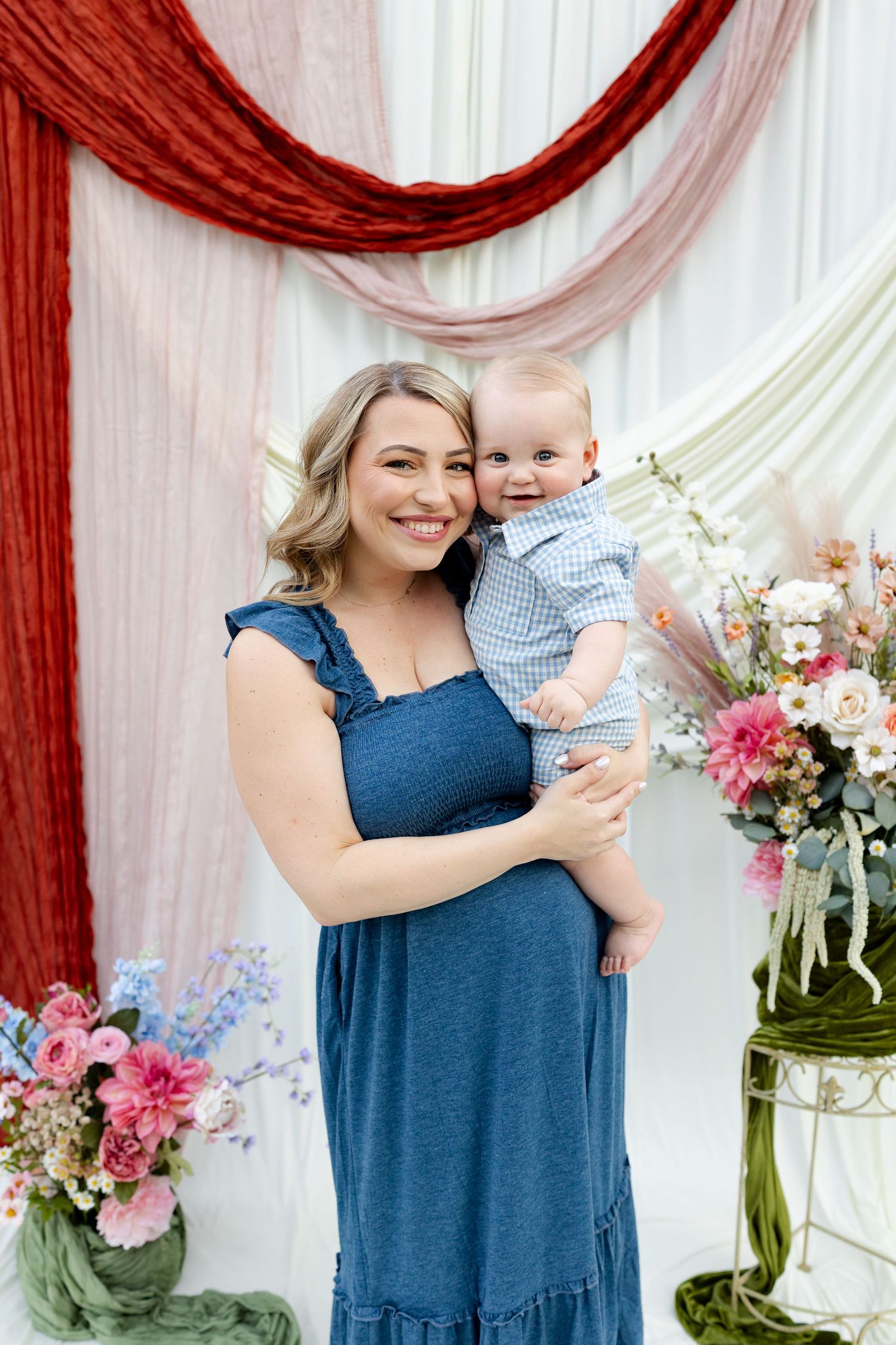 Woman in blue dress holds smiling baby boy in gingham outfit, posing before colorful floral backdrop.