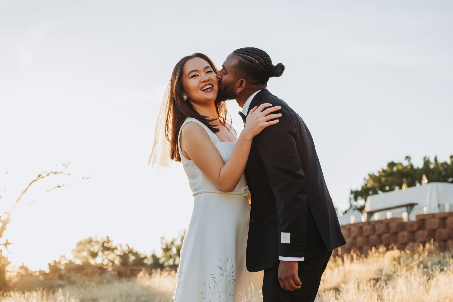 A joyful interracial couple shares a romantic kiss outdoors on their wedding day, bride in light blue dress.