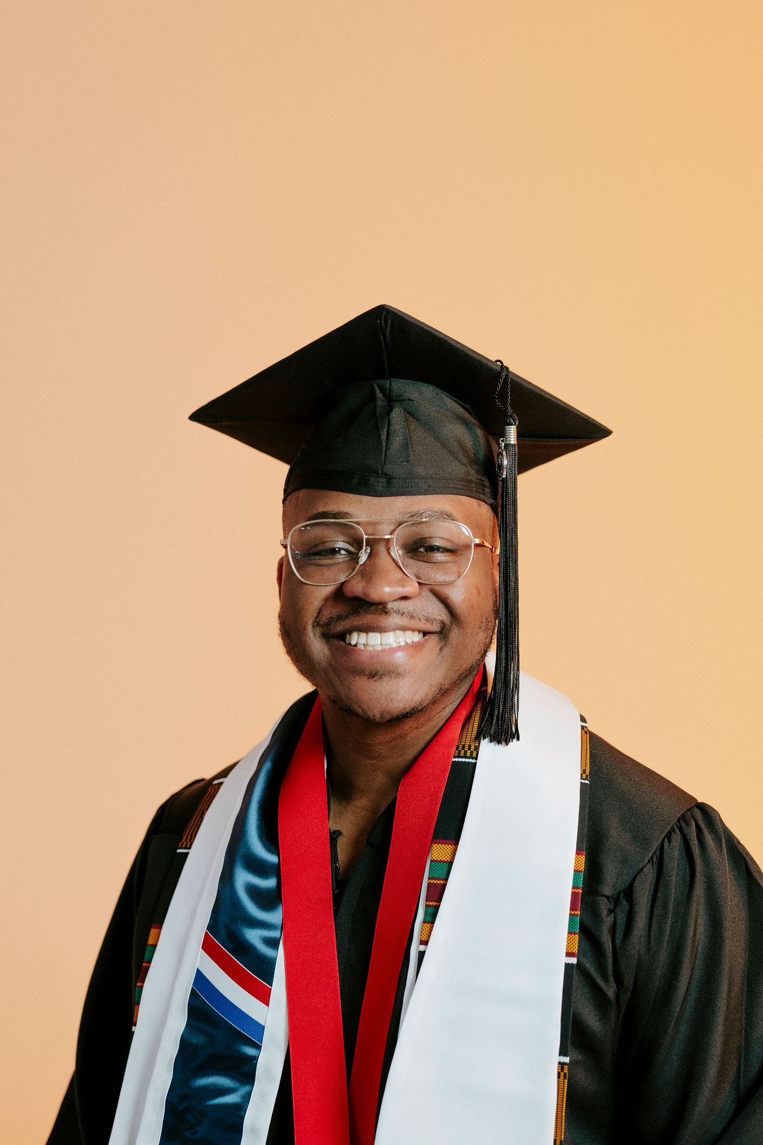 Graduate in black cap and gown with red stole stands with arms outstretched in celebratory poses against grey background.