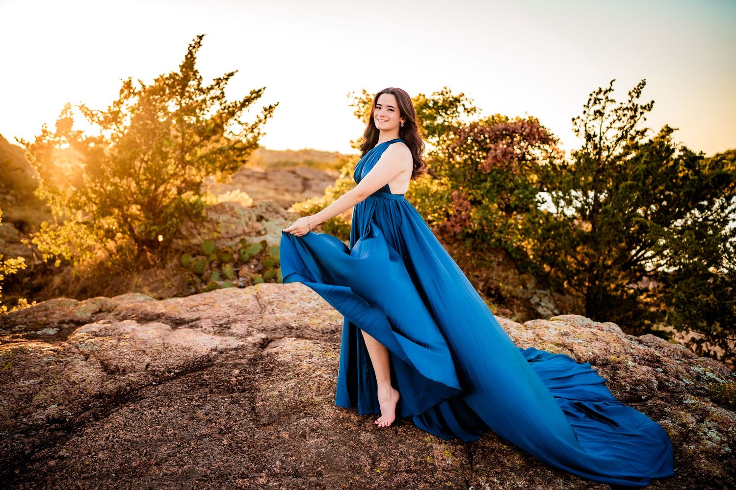 A woman in a flowing blue dress poses gracefully on rocky terrain during golden hour sunset.