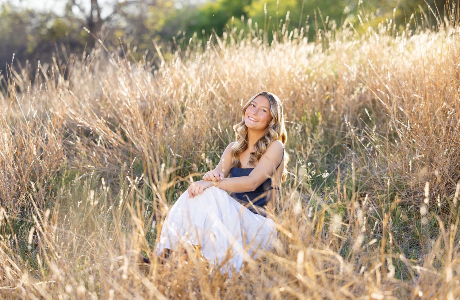 Young woman in navy dress leaning on white wooden fence in golden hour sunlight outdoors.