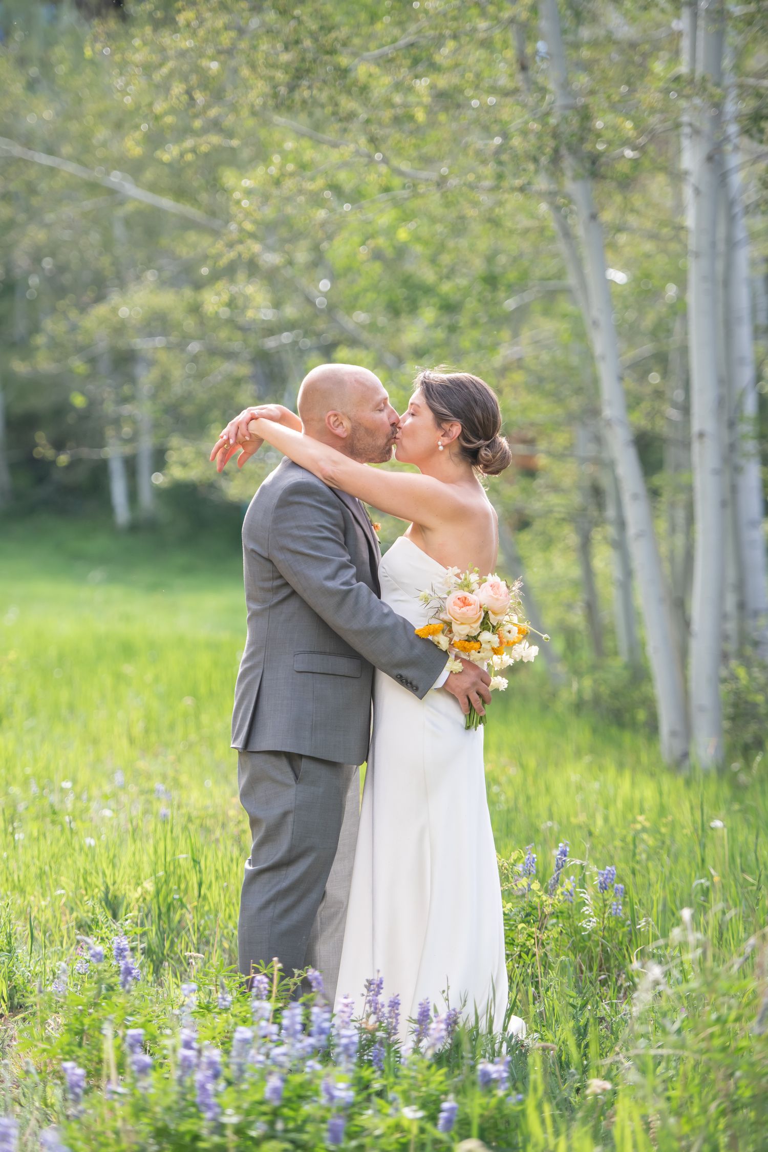 Bride and groom sharing a kiss in a wildflower meadow with aspen trees, bride holding a colorful bouquet.