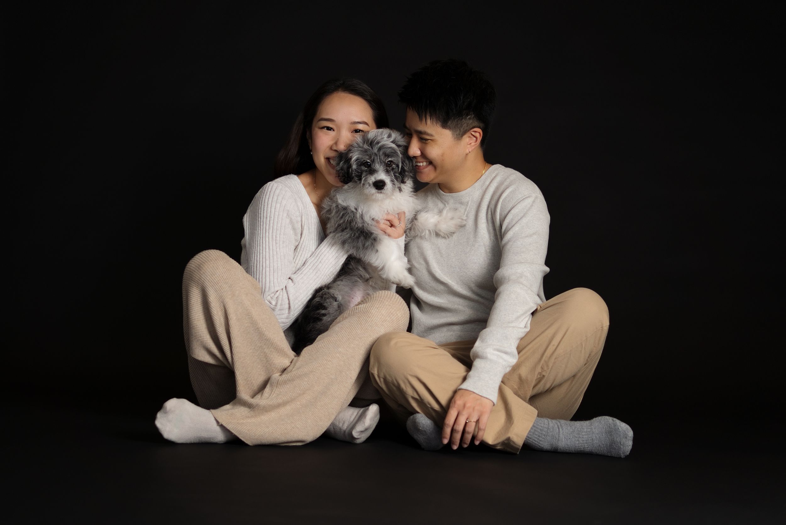 A smiling couple sits on a dark floor holding a fluffy gray and white dog between them. In home studio like photoshoot with a 14 week puppy