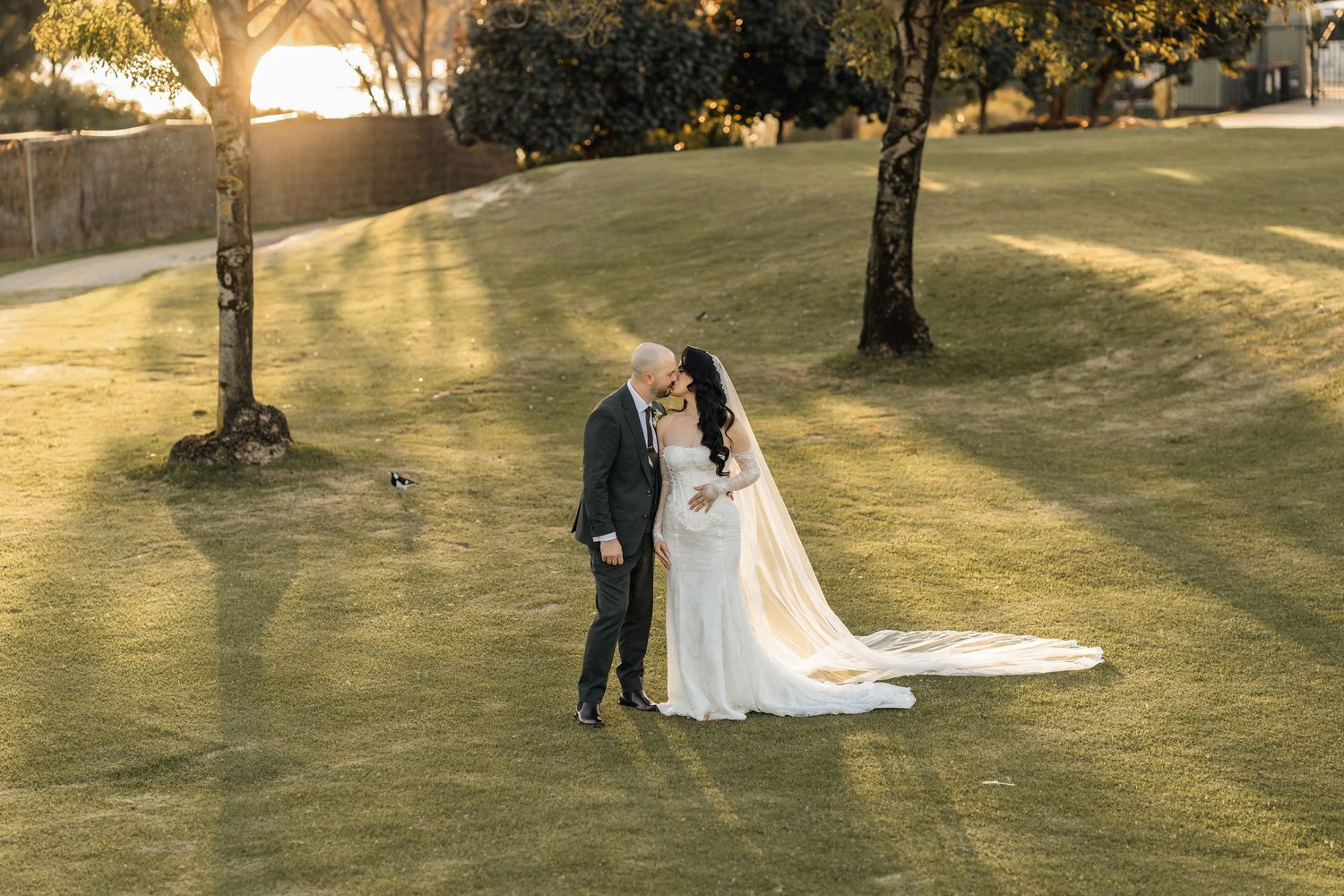 Bride and groom sharing a romantic kiss on a sunlit green lawn, with golden hour light glowing behind them.