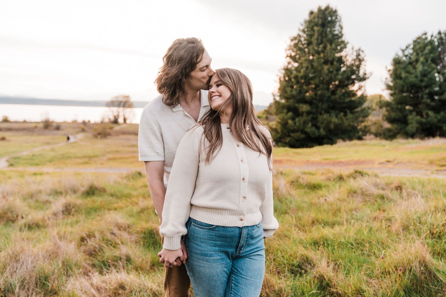 Happy couple holding hands in a green meadow, man kissing womans head, trees and lake in background.