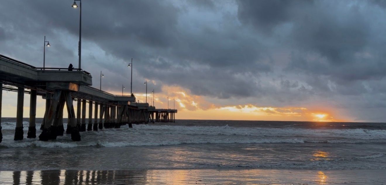 A dramatic stormy beach sunset with dark clouds and a wooden pier silhouetted against the orange sky.