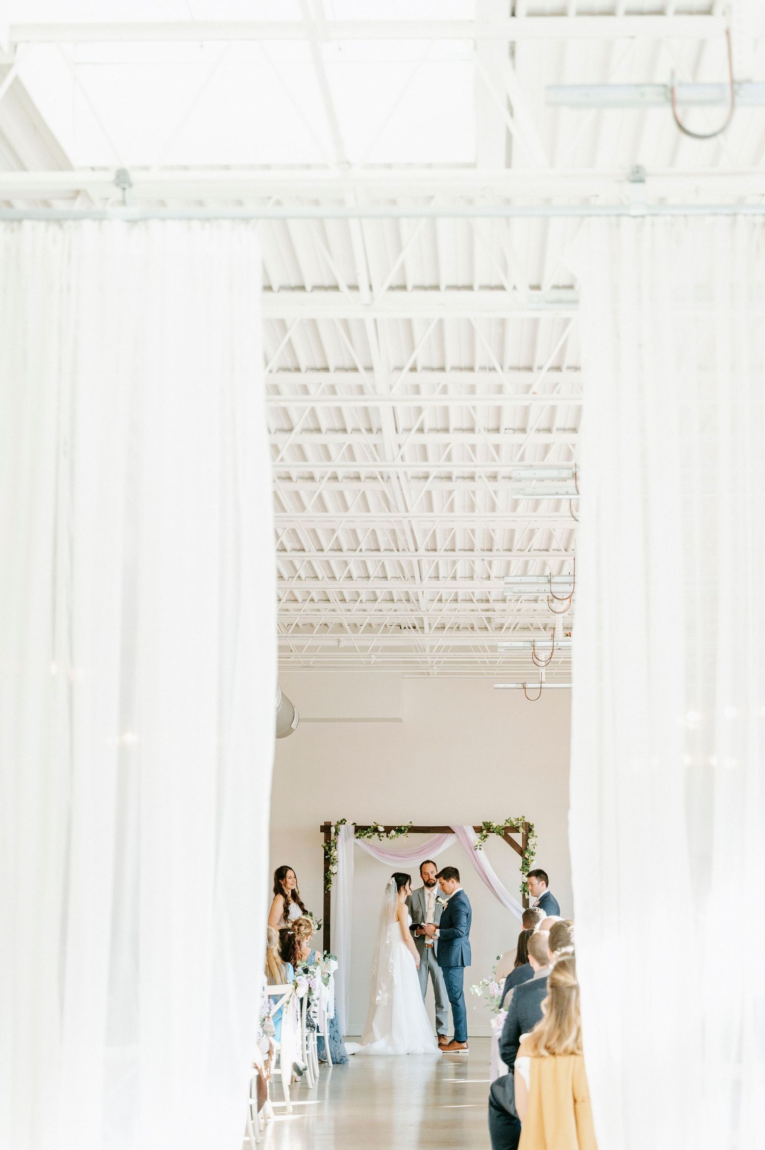 Elegant white wedding venue interior with sheer curtains and guests mingling in a bright modern space.