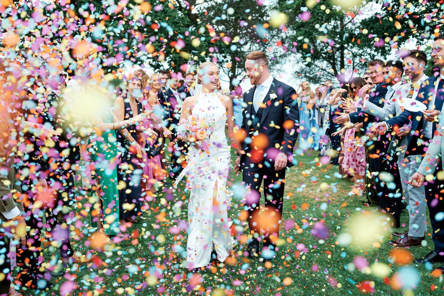 Bride and groom walking through a colourful confetti shower surrounded by wedding guests outdoors at Blake Hall