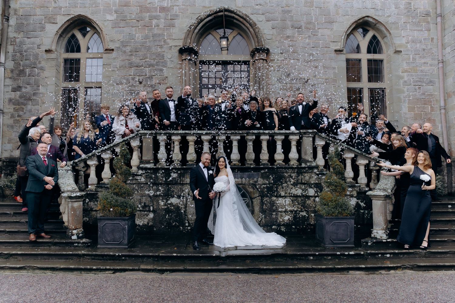 Wedding party poses for formal group photos along a stone balustrade at an elegant historic venue.
