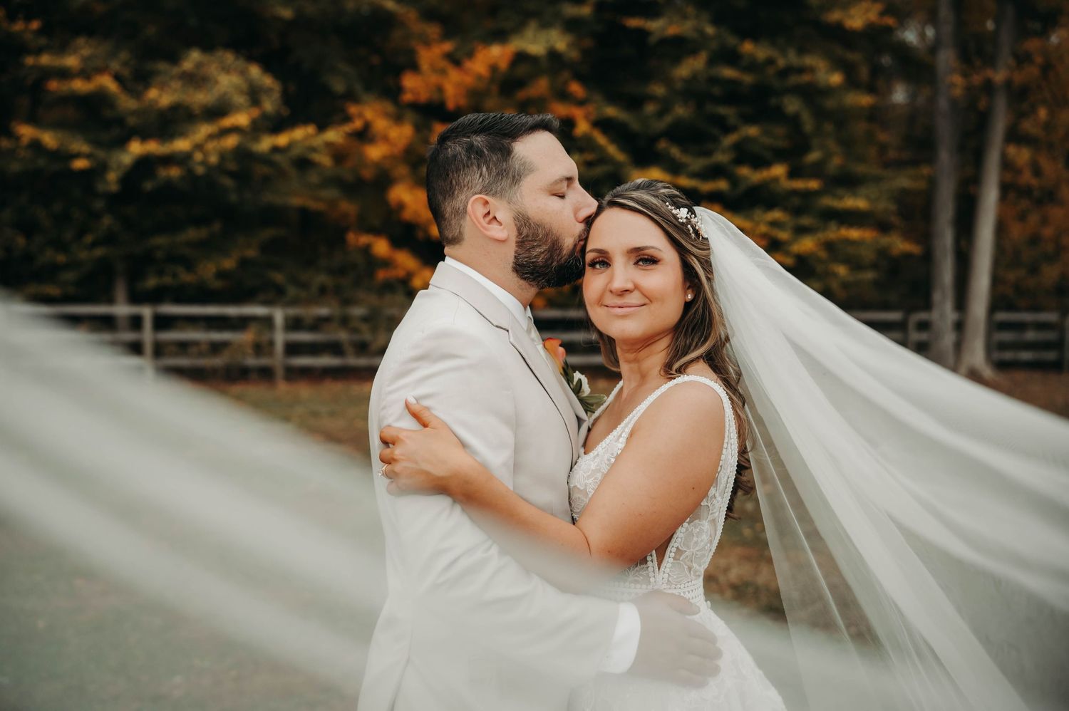 Bride and groom embracing outdoors with veil flowing amid autumn foliage backdrop.