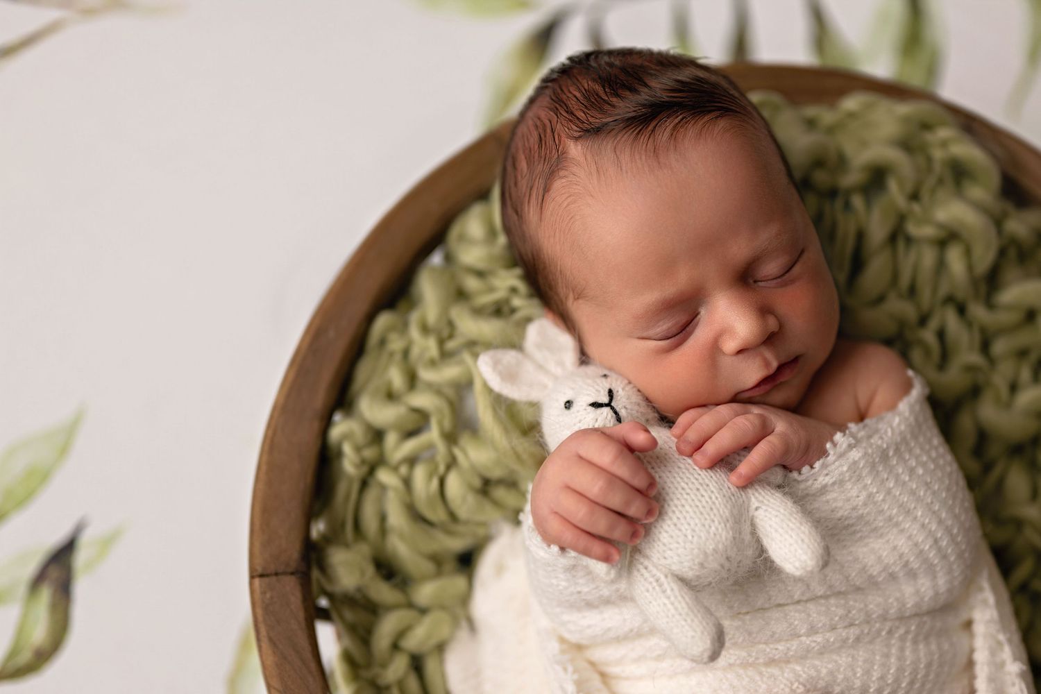 Sleeping newborn baby wrapped in white knit blanket holding a stuffed bunny toy in a wooden bowl.