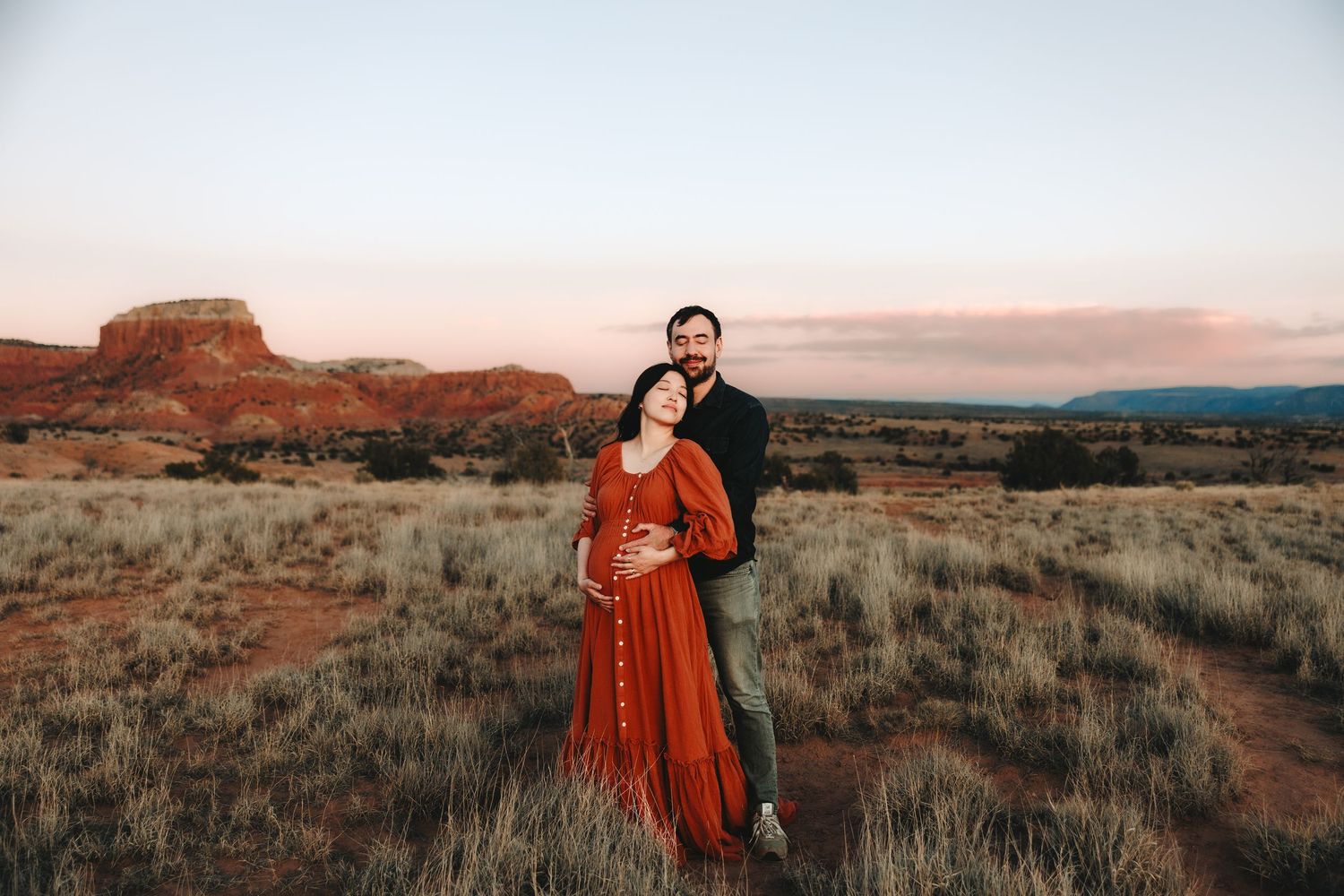 Couple embracing in red desert landscape at sunset during maternity photo session.