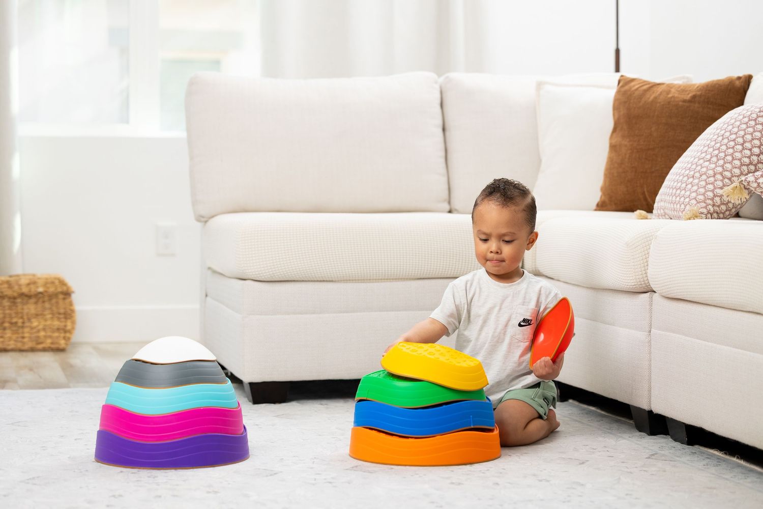 Toddler boy playing with colorful stacking sensory pads toy on white carpet next to a modern sofa. Image for Amazon listing.