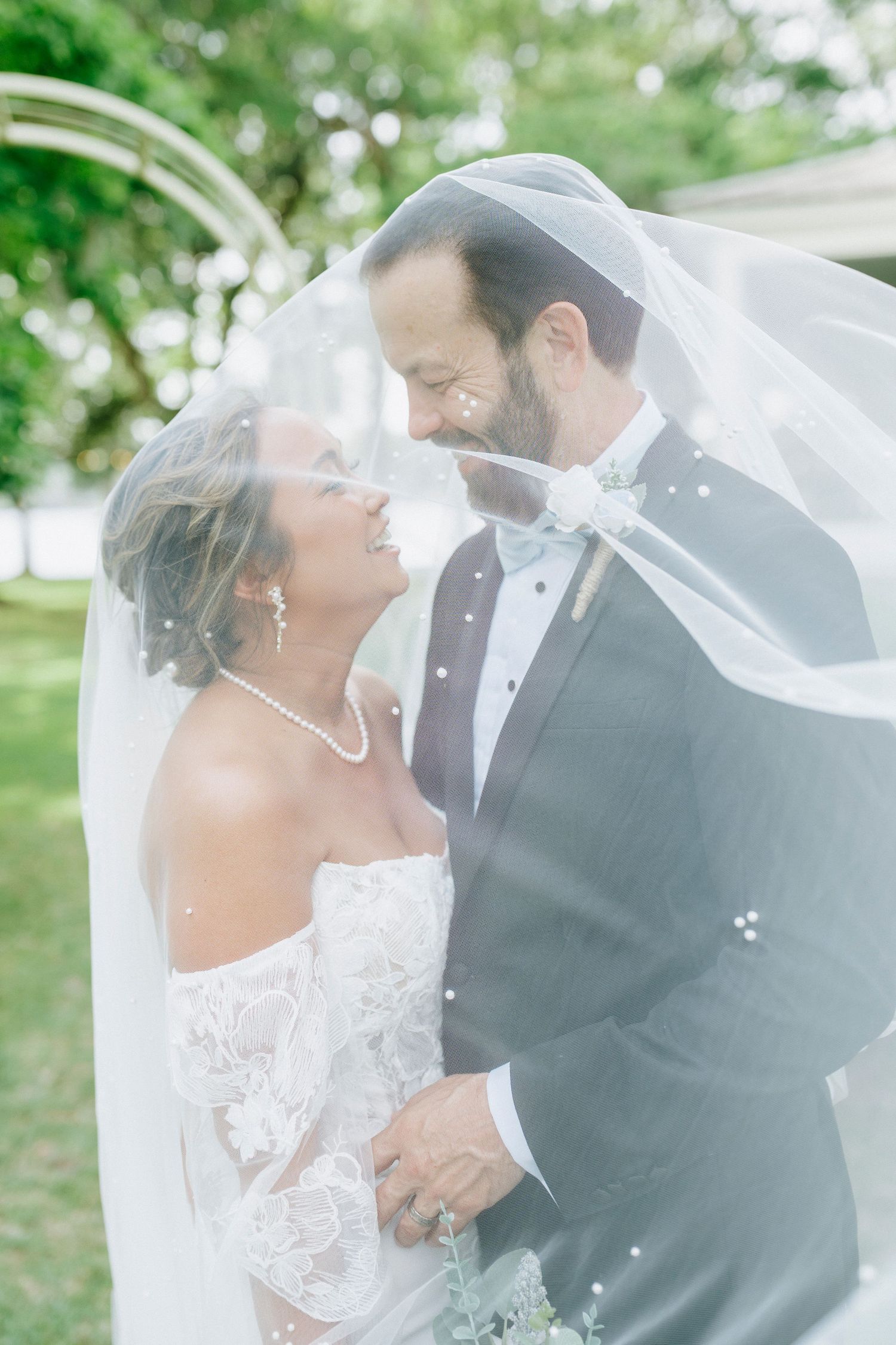 Bride and groom share a romantic moment under a flowing veil outdoors, smiling at each other in wedding attire.