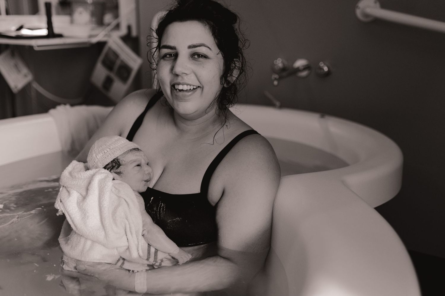 Black and white photo of a smiling mother holding her newborn baby in a birthing tub after water birth.