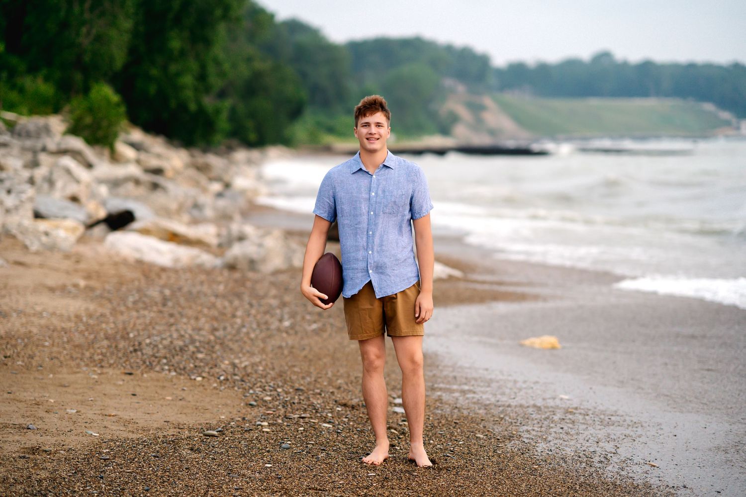Someone in a blue shirt and shorts stands barefoot on a rocky beach shoreline on a summer day.